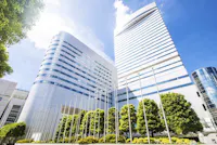 A modern urban scene featuring two tall, sleek office buildings with reflective glass surfaces under a bright blue sky. The foreground includes lush green trees and a series of flagpoles.