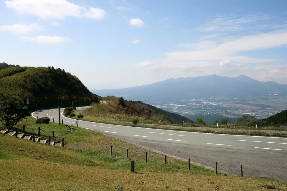 A winding mountain road curves through green hills under a partly cloudy sky. In the distance, a city is visible with a backdrop of expansive mountains. The landscape is lush and serene, highlighted by sunlight.
