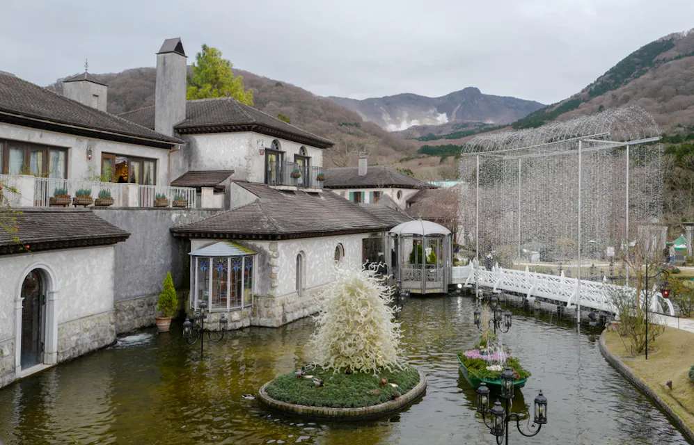 A picturesque scene of a European-style building by a serene pond with a white sculpture in the center. A small bridge crosses the pond, and mountains loom in the background under a cloudy sky.
