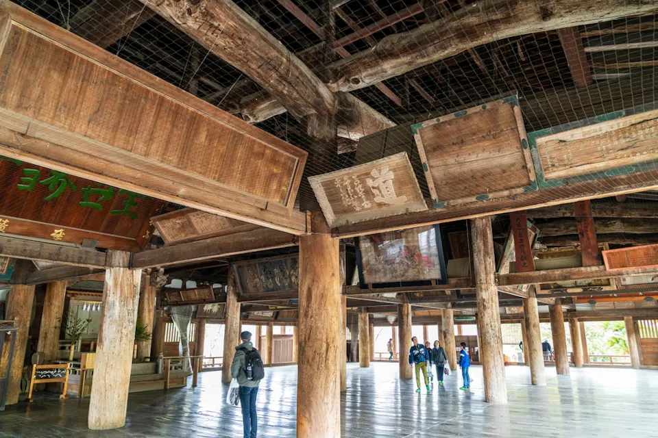 Interior of a traditional wooden temple with large wooden beams and pillars. Several people are walking around, and various wooden plaques with inscriptions are displayed overhead. The floor is made of polished wood.