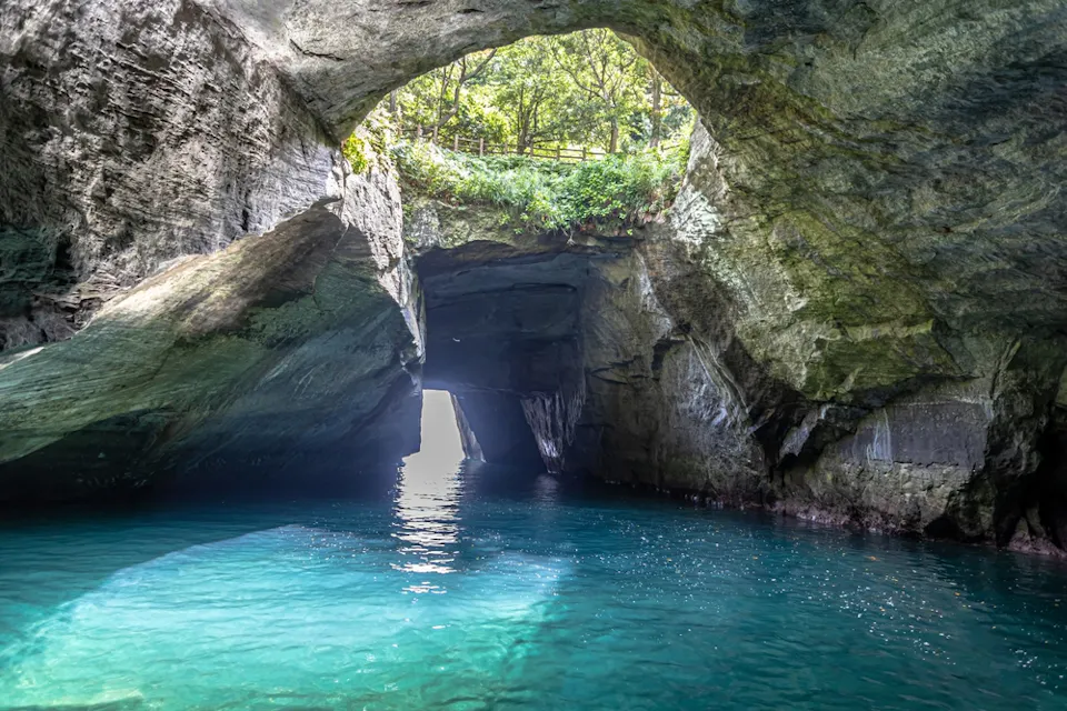 A scenic view of a cave with turquoise water flowing through it. Sunlight illuminates the entrance, highlighting the rugged rock walls and the greenery above. The cave opens to a bright, sunlit area in the distance.
