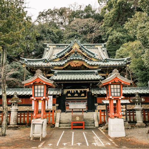 Shizuoka Sengen Shrine A traditional Japanese shrine entrance, featuring a grand wooden gate with intricate carvings and green-tiled roof. Red and white lanterns stand on either side. Lush green trees surround the serene setting, along with a stone tablet inscribed in Japanese.