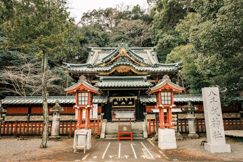 A traditional Japanese shrine entrance, featuring a grand wooden gate with intricate carvings and green-tiled roof. Red and white lanterns stand on either side. Lush green trees surround the serene setting, along with a stone tablet inscribed in Japanese.