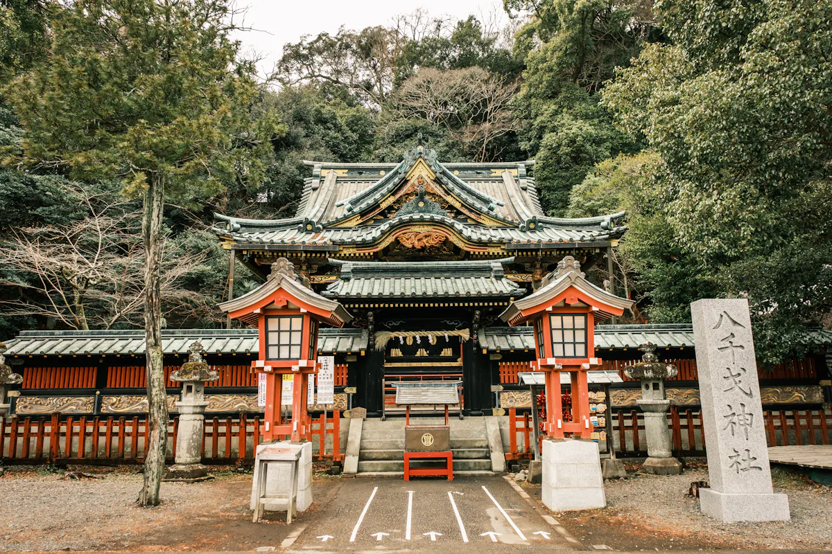 Shizuoka Sengen Shrine A traditional Japanese shrine entrance, featuring a grand wooden gate with intricate carvings and green-tiled roof. Red and white lanterns stand on either side. Lush green trees surround the serene setting, along with a stone tablet inscribed in Japanese.