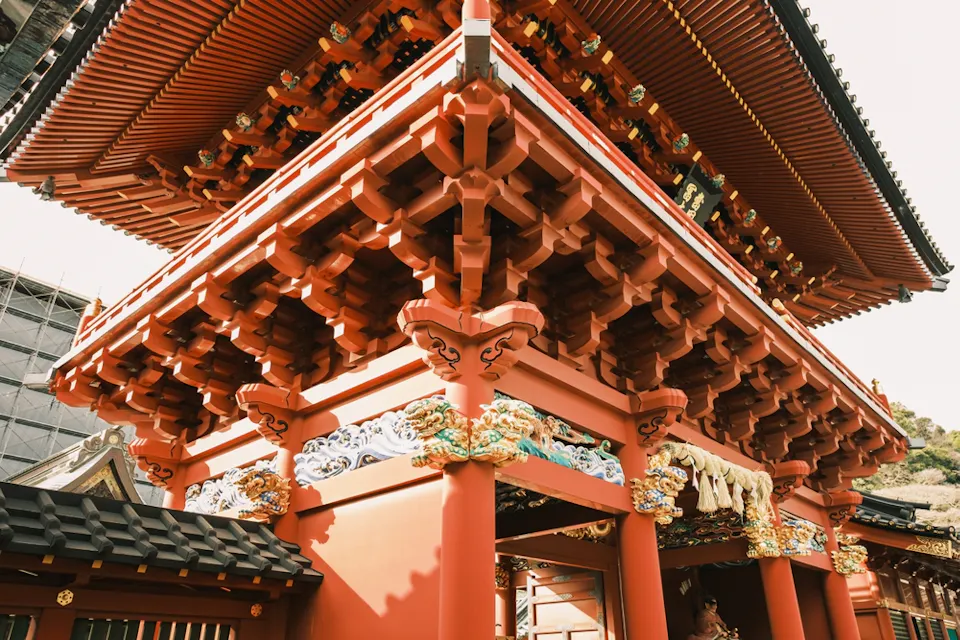 A close-up view of a traditional Japanese temple gate, featuring red wooden beams and intricate carvings. Gold and colorful embellishments decorate the structure, with a clear blue sky in the background.