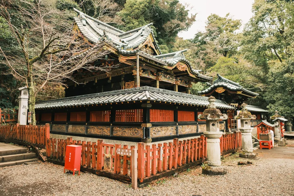 An ancient Japanese shrine surrounded by trees, featuring traditional wooden architecture with ornate roof details. Stone lanterns line the gravel path, and a red fence encircles the building, creating a serene and historic atmosphere.