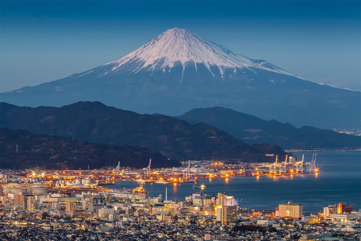 A nighttime view of a well-lit coastal city with a bustling harbor, set against the backdrop of a towering snow-capped mountain under a clear sky.