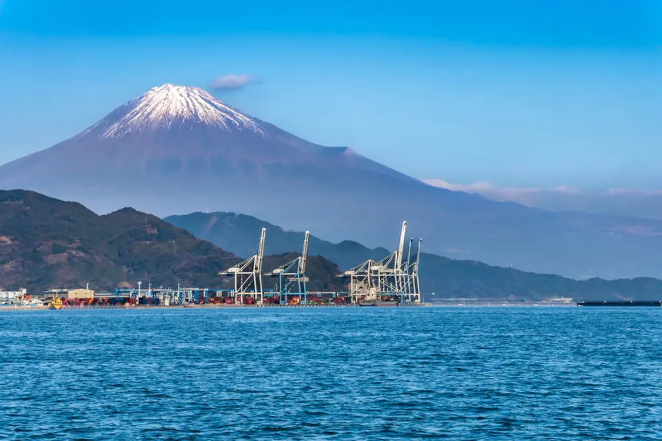 Shimizu Port A scenic view of Mount Fuji with a snow-capped peak in the background, overlooking a calm body of water. In the foreground, there is a harbor with cranes and industrial structures against a clear blue sky.