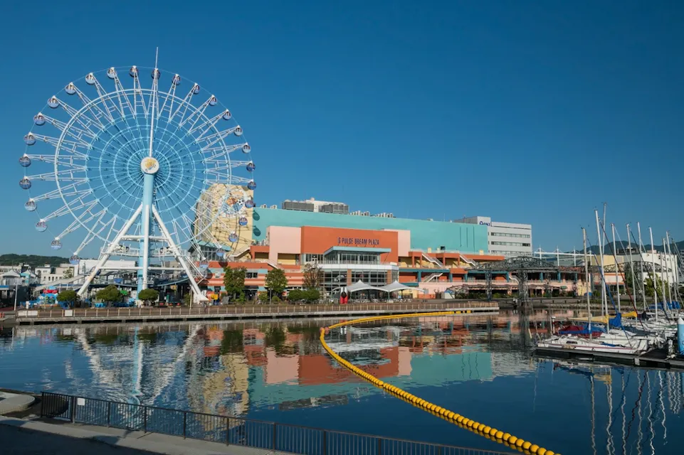 S-Pulse Dream Plaza A vibrant waterfront scene with a large Ferris wheel, colorful buildings including a blue and orange complex labeled "Space World Park," and boats docked along the pier. The clear blue sky is reflected in the calm water.
