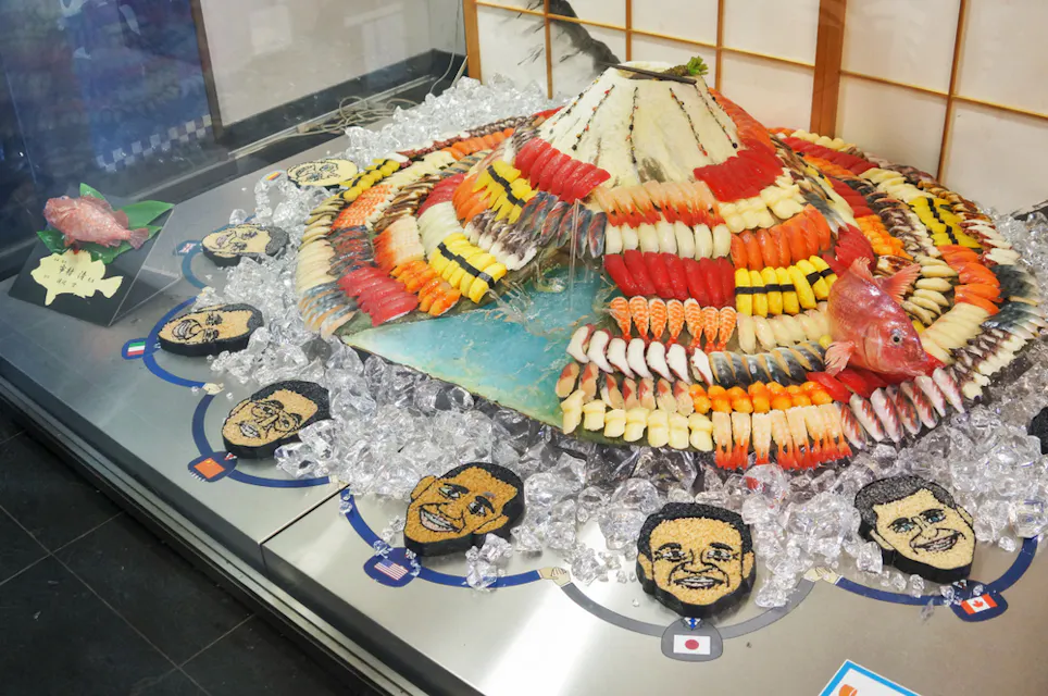 Sushi Museum A display of a volcano-shaped sushi platter, surrounded by ice, with an assortment of fish and faces made of food depicting various people. The arrangement includes vibrant colors with various sushi types and a fish placed on top.