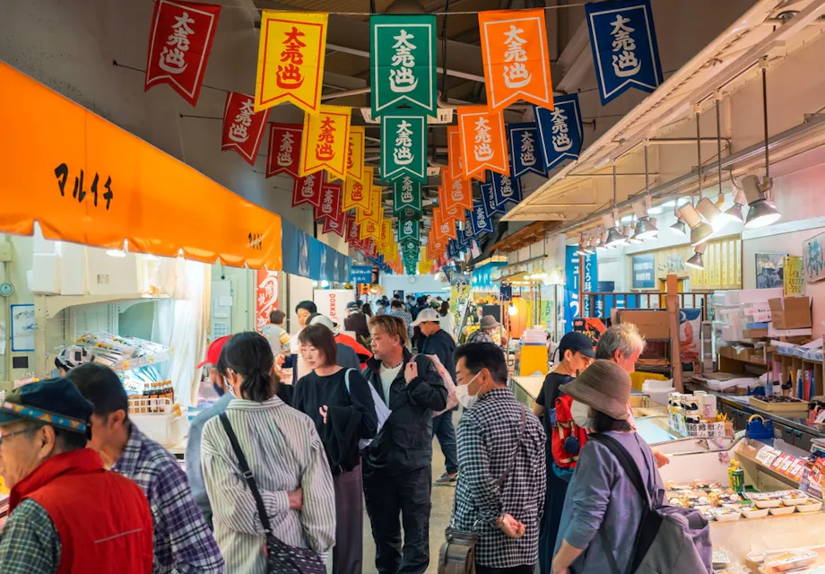 Kashi no Ichi Market A bustling indoor market scene with colorful banners hanging from the ceiling. Shoppers browse stalls offering various products. The atmosphere is lively with people engaged in conversation and exploring the market goods.