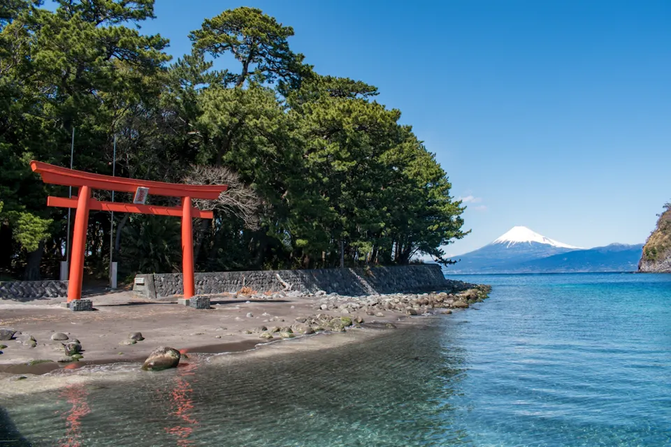 A scenic view of a red torii gate on a beach with clear blue water. Lush green trees line the shore, and Mount Fuji is visible in the background under a clear blue sky.
