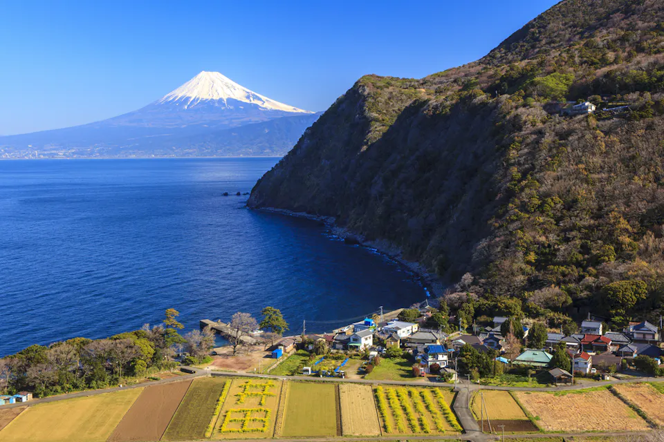 A scenic landscape with Mount Fuji in the background under a clear blue sky. The foreground features a coastal village with fields and houses, set against a blue sea and lush hillside.