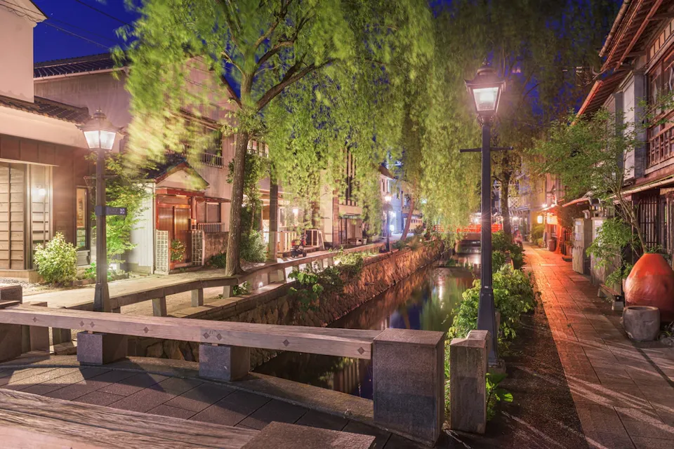 A serene, lantern-lit street at night in Japan features a narrow canal lined with trees and traditional buildings. The scene captures the peaceful ambiance with a blend of greenery and illuminated architecture.