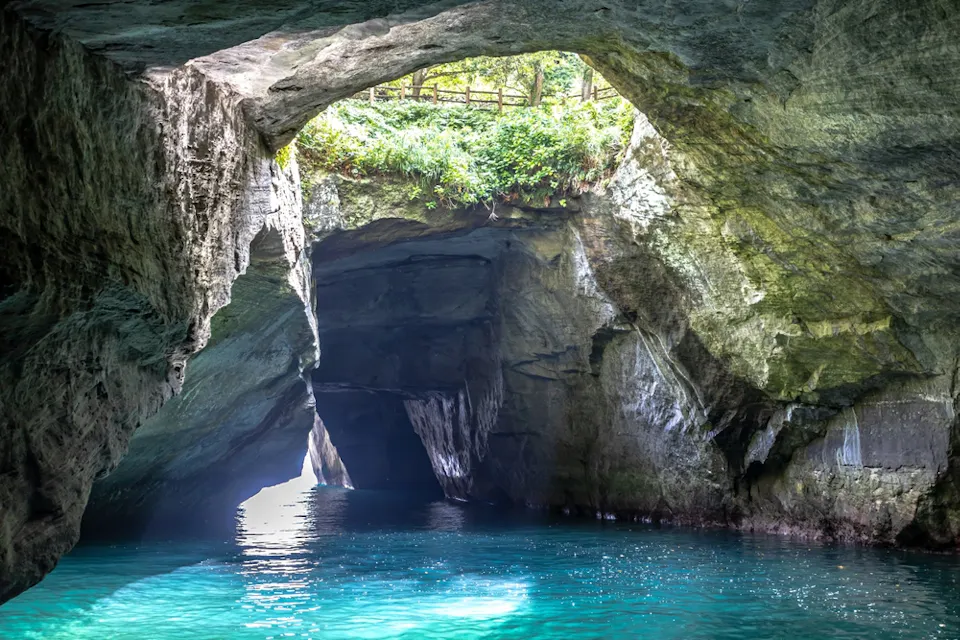 A scenic view of a cave with turquoise water inside. Sunlight filters through the entrance, illuminating the rock formations and highlighting the lush greenery at the top.