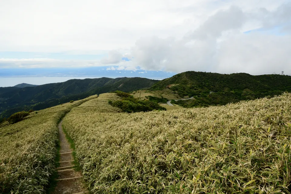 A narrow path winds through lush, grassy hills under a partly cloudy sky. The landscape features rolling hills and distant mountains, while the horizon reveals a faint glimpse of the sea. The scene evokes a sense of tranquility and open space.