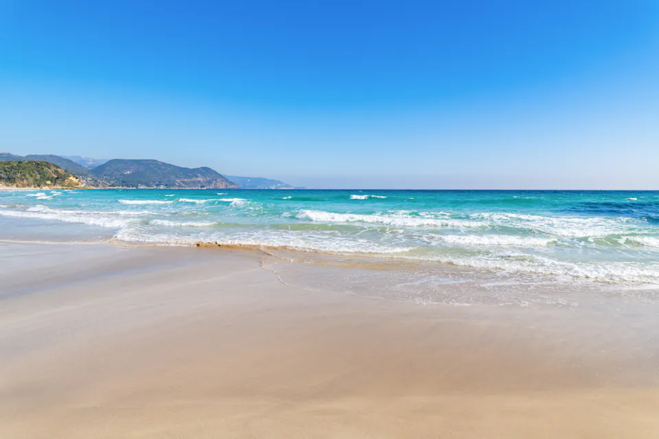 A serene beach scene with gentle waves lapping at the shore under a clear blue sky. The turquoise sea stretches out to meet the horizon, with distant hills visible on the left side. The sandy foreground glistens in the sunlight.