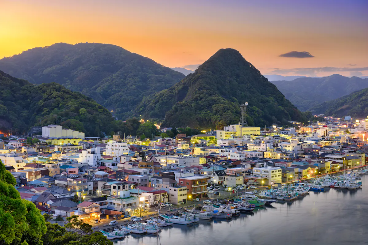 A coastal town nestled between lush green hills at sunset, with buildings illuminated by streetlights. Boats are docked along a calm harbor, and the sky transitions from orange to deep blue above the silhouetted mountains.