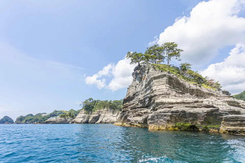 Rocky cliffs with layers of sediment rise dramatically from the blue ocean, dotted with green trees on top. The sky is clear with a few fluffy clouds, creating a picturesque coastal scene.