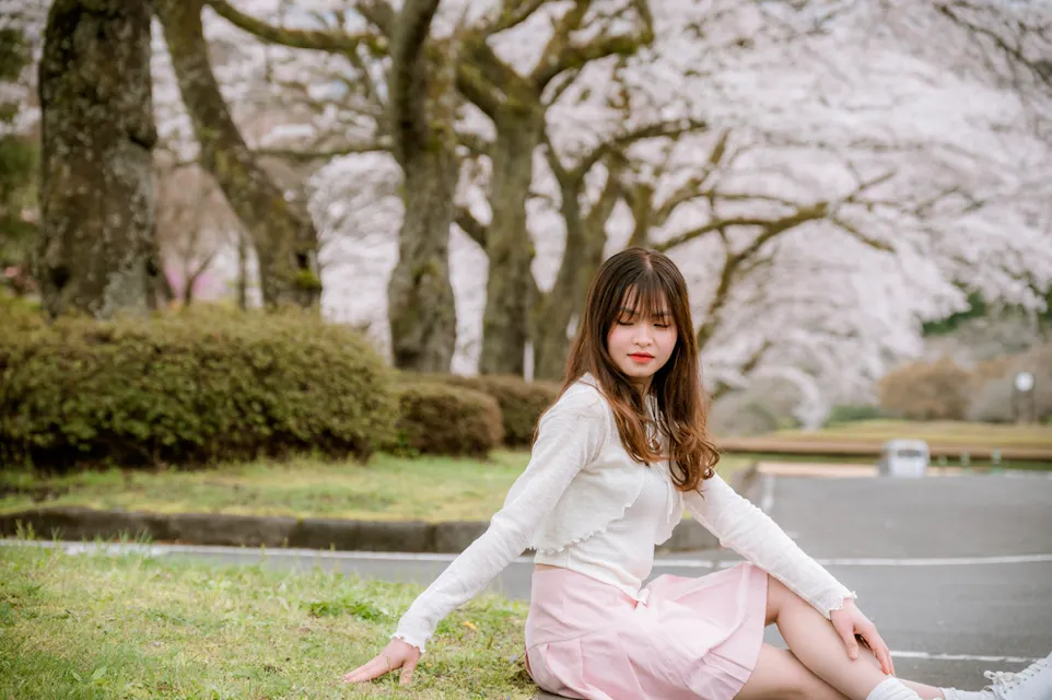 A woman sits on the grass in a park, surrounded by blooming cherry blossom trees. She wears a light-colored outfit and looks down, with a serene expression. The background features rows of trees lining a pathway.