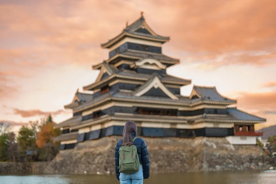 A person with a green backpack stands in front of a traditional Japanese castle with multiple tiers and black and white architecture. The sky is orange and pink, suggesting a sunset or sunrise. The foreground includes a reflective body of water.