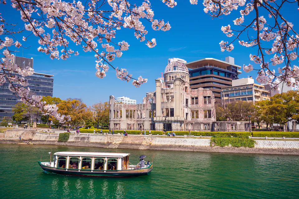 A boat floats on the river with cherry blossoms in the foreground. The Hiroshima Peace Memorial, surrounded by trees and modern buildings, is visible under a clear blue sky.