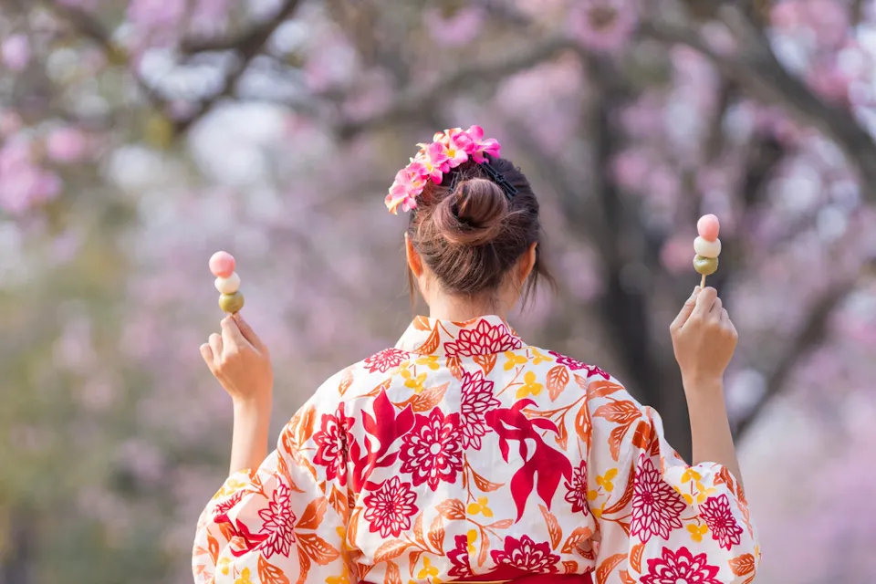 A person wearing a floral kimono and a flower hair accessory holds two dango skewers. They are standing outdoors with blurred cherry blossoms in the background.