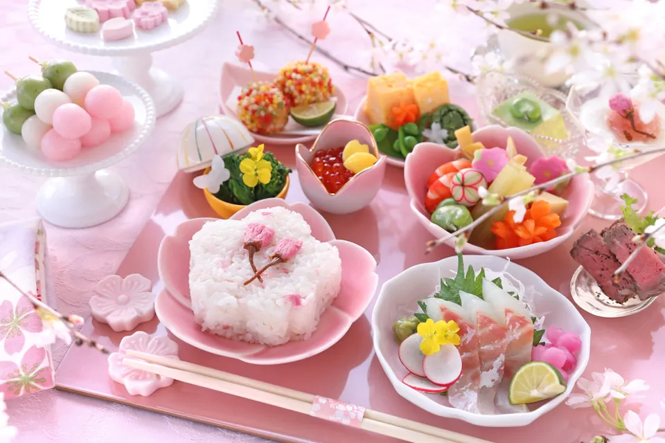 A beautifully arranged Japanese meal set on a pink table. It includes rice shaped like a flower, various colorful sushi, vegetables, and desserts. Surrounded by cherry blossom decorations and chopsticks in the foreground.