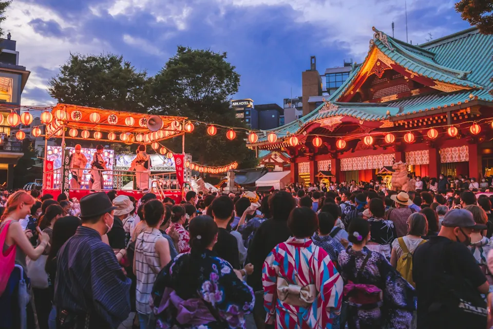 A vibrant festival scene with a crowd gathered in front of a traditional Japanese temple. Participants wearing colorful yukatas watch dancers on a stage adorned with red lanterns. The sky is a mix of blue and clouds, creating a festive atmosphere.