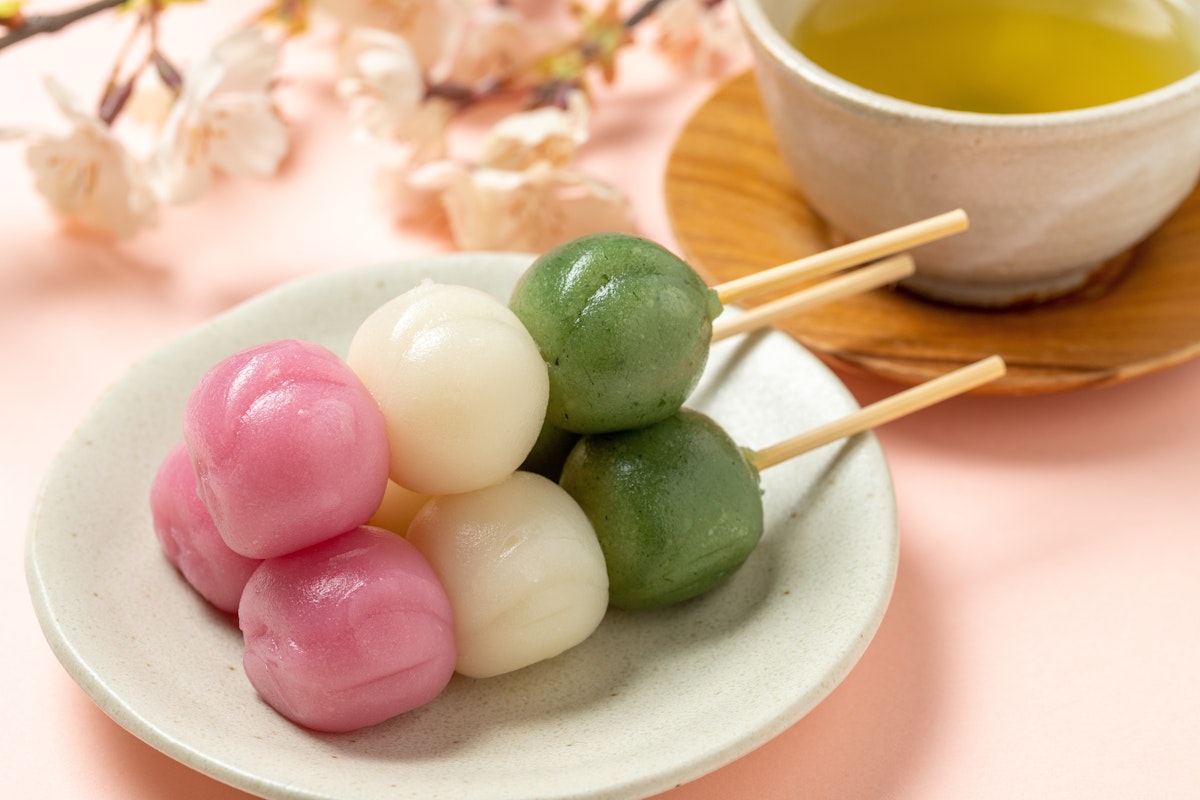 Japanese Spring Food A plate of colorful dango skewers with pink, white, and green mochi balls sits on a light pink surface. In the background, a cup of green tea and cherry blossoms are visible, adding to the traditional Japanese setting.