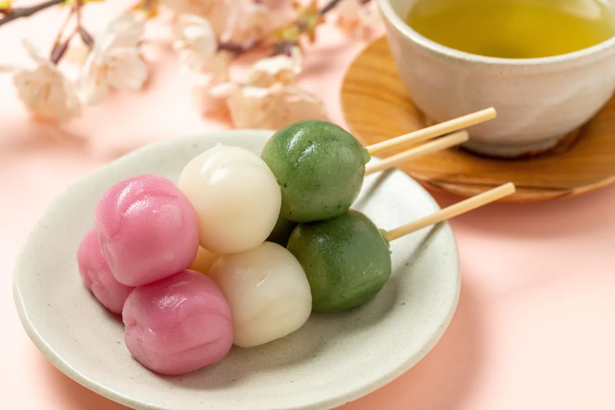 A plate of colorful dango skewers with pink, white, and green mochi balls sits on a light pink surface. In the background, a cup of green tea and cherry blossoms are visible, adding to the traditional Japanese setting.