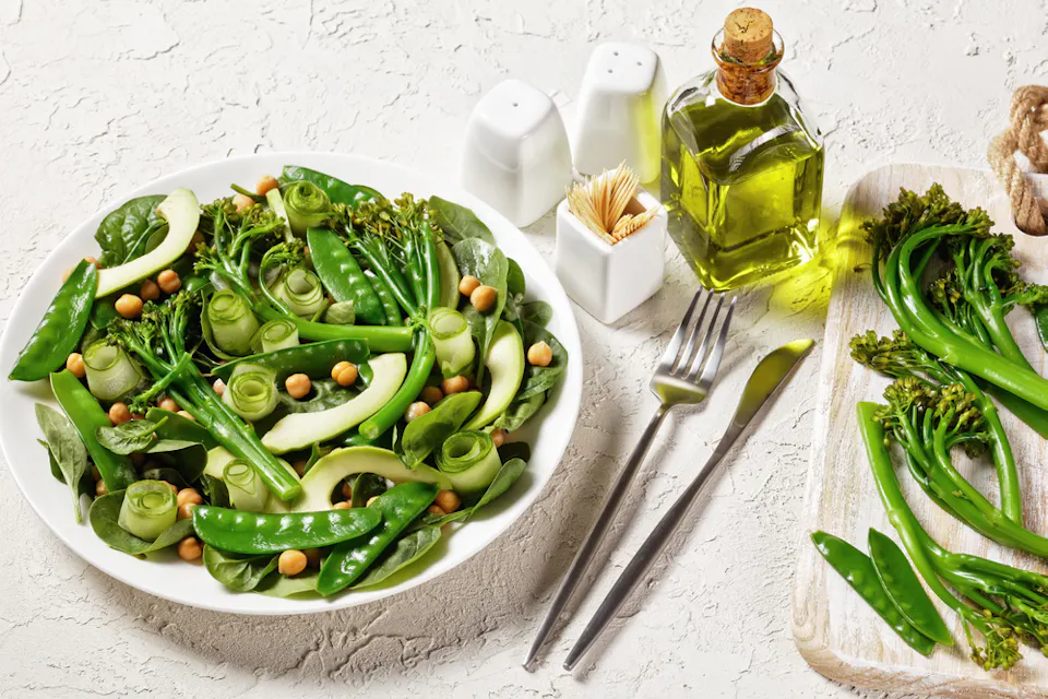 A fresh green salad with snap peas, chickpeas, spinach, cucumber ribbons, and broccolini is arranged on a white plate. Olive oil, salt, pepper shakers, and cutlery are set beside it on a textured white surface.