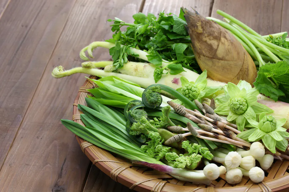 A bamboo basket filled with a variety of fresh green vegetables, including leafy greens, fiddlehead ferns, and bamboo shoots, placed on a wooden surface.