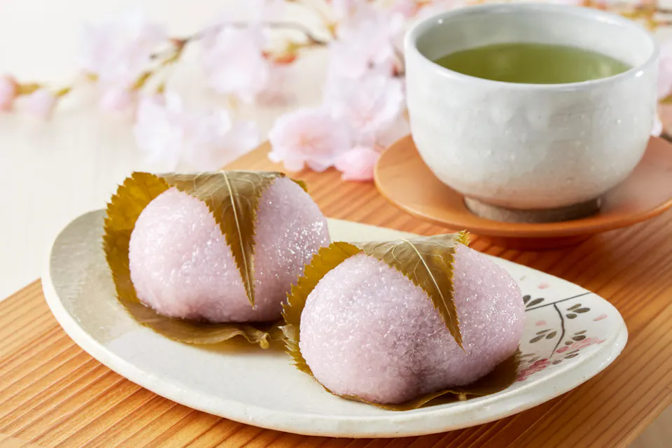 Two pink sakura mochi wrapped in cherry blossom leaves on a decorative plate, accompanied by a cup of green tea. Delicate cherry blossoms adorn the background, adding a touch of elegance to the setting.