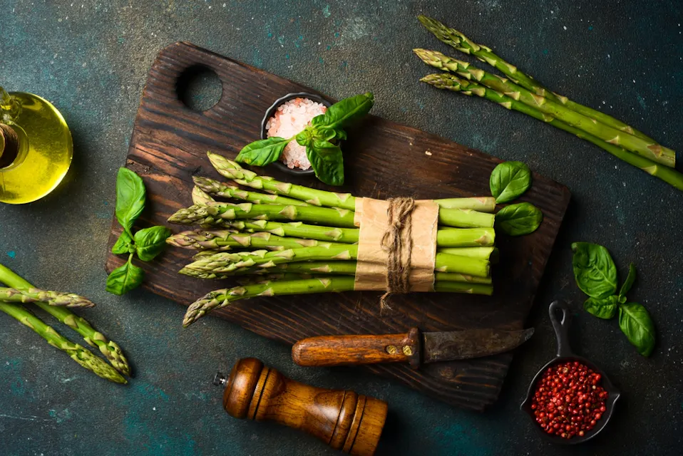 A bunch of fresh asparagus tied with twine rests on a dark wooden cutting board with pink salt. Surrounding items include basil, a small bottle of olive oil, a pepper mill, a knife, and a small dish of red peppercorns on a rustic surface.