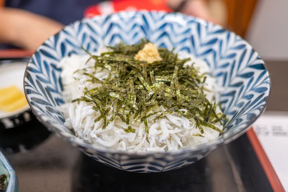 A patterned blue and white bowl filled with thin, white noodles topped with shredded seaweed and a small dollop of ginger. The dish is placed on a black tray with another small plate visible in the background.