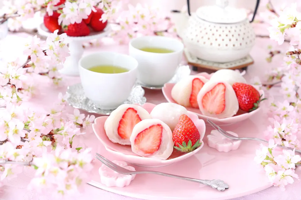 A beautifully arranged scene with plates of strawberry mochi surrounded by delicate cherry blossoms. Two cups of green tea sit beside a white teapot, all set on a pastel pink table. The setting is serene and elegant.