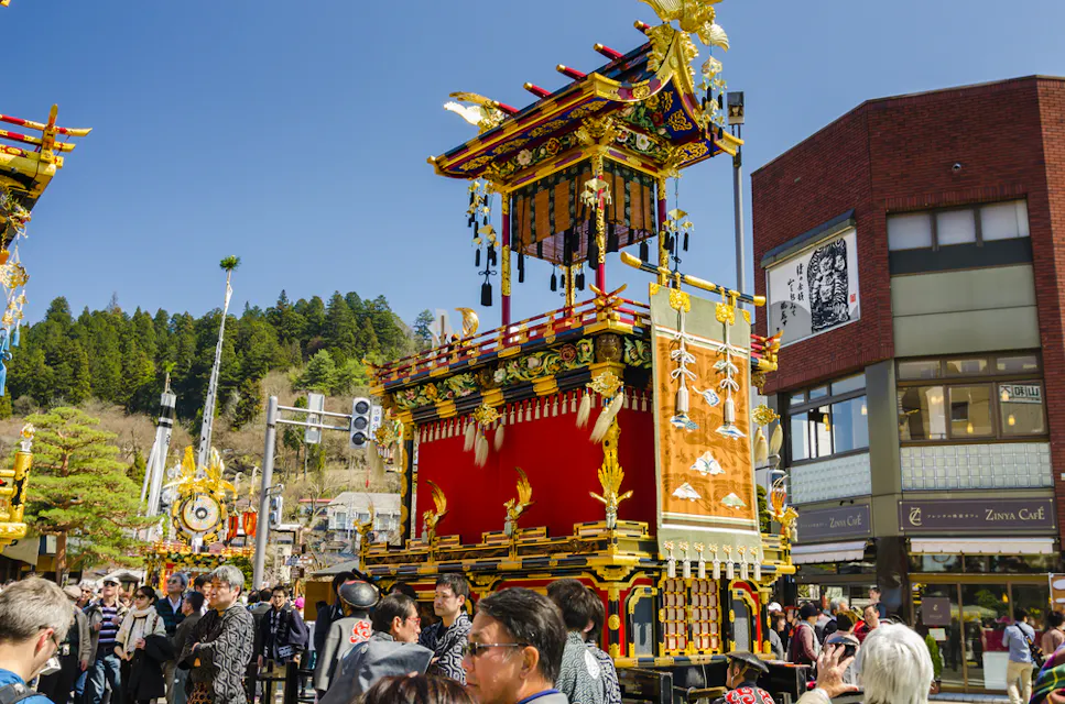 A vibrant street scene depicts a traditional Japanese festival with an ornately decorated float adorned with gold and red details. People in festive clothing gather around, enjoying the sunny day amidst a backdrop of trees and modern buildings.