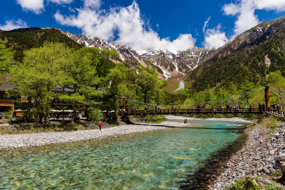A clear turquoise river flows through a rocky bed, surrounded by lush green trees. A wooden bridge with people crosses the river. In the background, snow-capped mountains rise under a bright blue sky with scattered clouds.