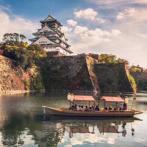 Osaka, Japan A traditional boat filled with people floats on a calm moat surrounding Osaka Castle, Japan. The castle stands majestically with its stone walls and elegant roof, under a partly cloudy sky. Lush trees with autumn foliage add color to the scene.