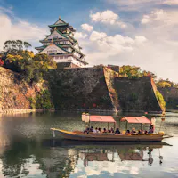 Osaka, Japan A traditional boat filled with people floats on a calm moat surrounding Osaka Castle, Japan. The castle stands majestically with its stone walls and elegant roof, under a partly cloudy sky. Lush trees with autumn foliage add color to the scene.