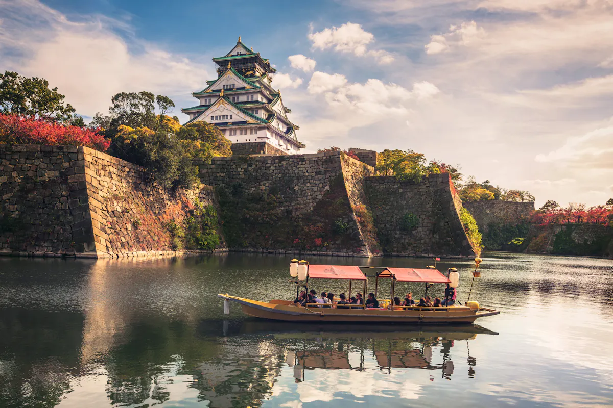 Osaka, Japan A traditional boat filled with people floats on a calm moat surrounding Osaka Castle, Japan. The castle stands majestically with its stone walls and elegant roof, under a partly cloudy sky. Lush trees with autumn foliage add color to the scene.