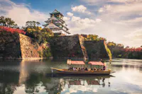 A traditional boat filled with people floats on a calm moat surrounding Osaka Castle, Japan. The castle stands majestically with its stone walls and elegant roof, under a partly cloudy sky. Lush trees with autumn foliage add color to the scene.