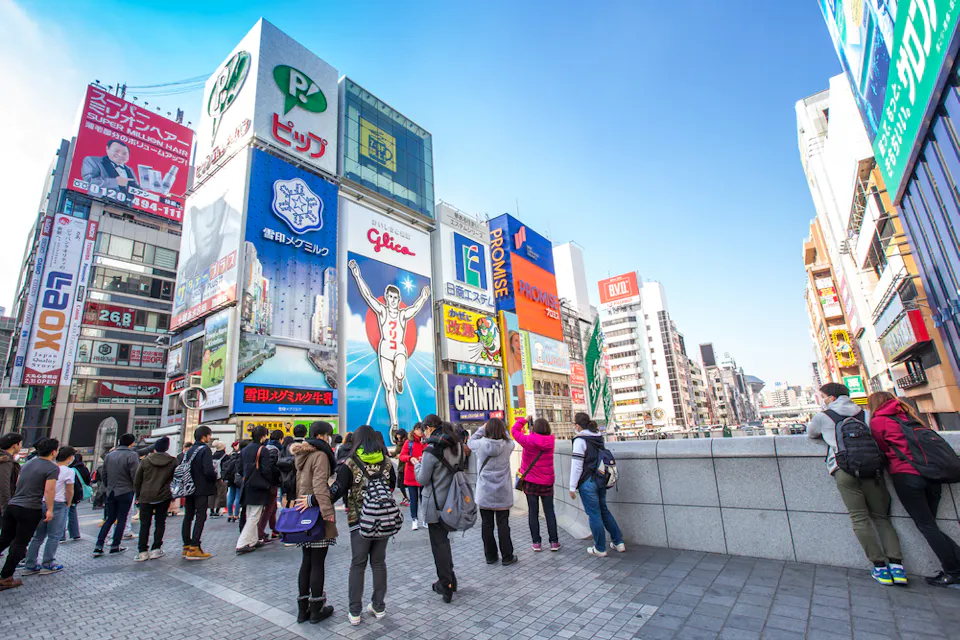 A bustling street scene in Osaka, Japan, with people gathered by a river. The area is filled with bright, colorful billboards, including a famous running man sign. The sky is clear and blue, adding vibrancy to the lively atmosphere.