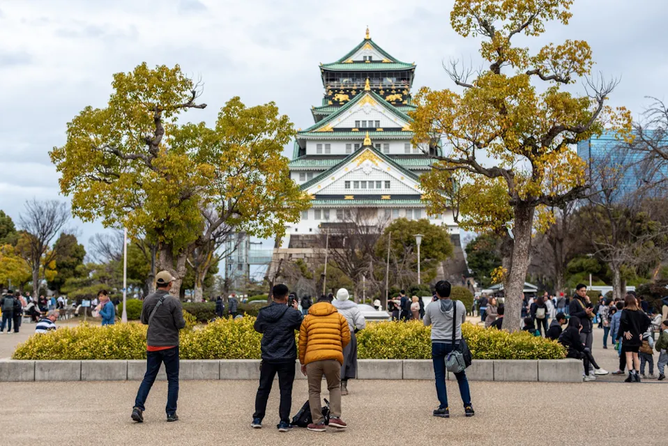 A group of people take photos in front of Osaka Castle, surrounded by vibrant trees and a gathering of visitors. The sky is overcast, adding a soft light to the historic scene.