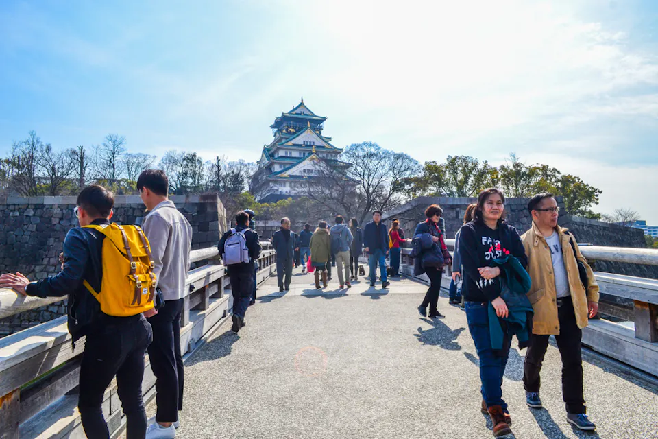People walking on a bridge towards Osaka Castle on a sunny day. The historic castle is visible in the background, surrounded by leafless trees. Visitors are casually dressed, and the atmosphere is lively.