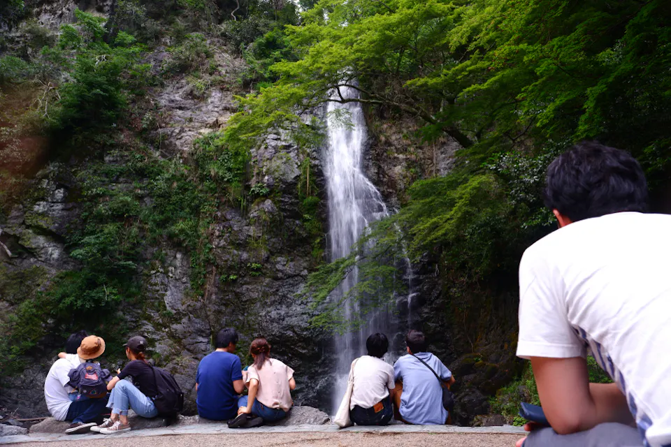 A group of people sitting on a rock, facing a tall waterfall cascading down a rocky cliff surrounded by lush green trees. The foreground features a person in a white shirt, back turned, observing the scene.