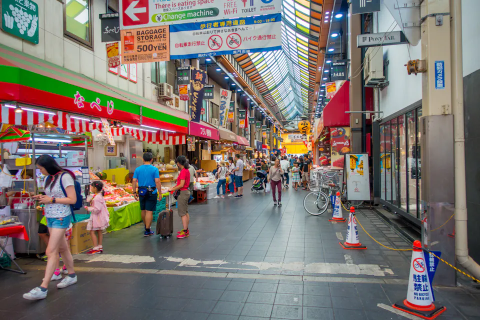 A vibrant indoor market scene with colorful storefronts and busy shoppers. Numerous signs hang from the ceiling, and bicycles are parked to the side. People of all ages are exploring the stalls in a lively atmosphere.