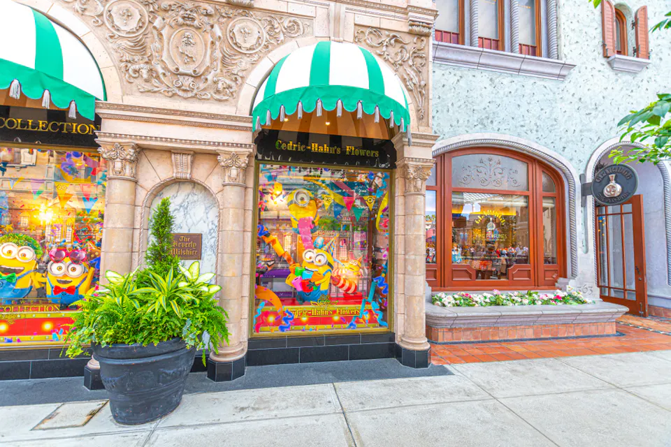 A whimsical storefront with colorful Minion-themed displays and green-striped awnings. The decorative facade features intricate carvings, and a potted plant sits outside. A neighboring shop has a large arched window with floral decorations.