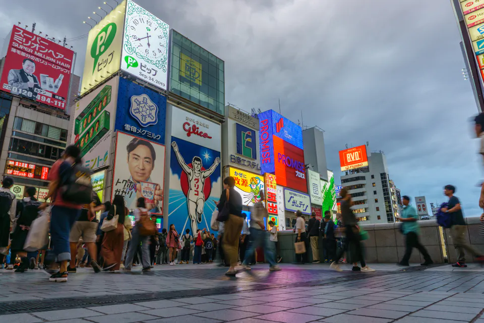 Crowded street in Dotonbori, Osaka, Japan, with vibrant neon signs and billboards, including the iconic Glico Running Man. People are walking along the street, creating a lively and bustling atmosphere under a cloudy sky.